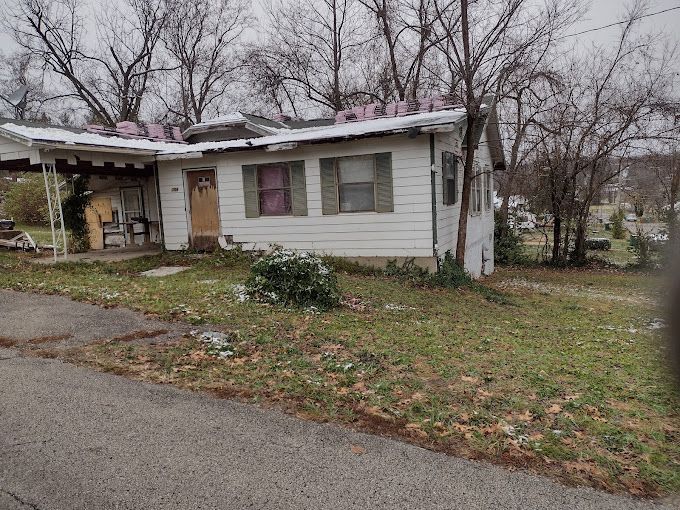 A rundown, white house with a damaged roof sits on a grassy lot on a gray day.