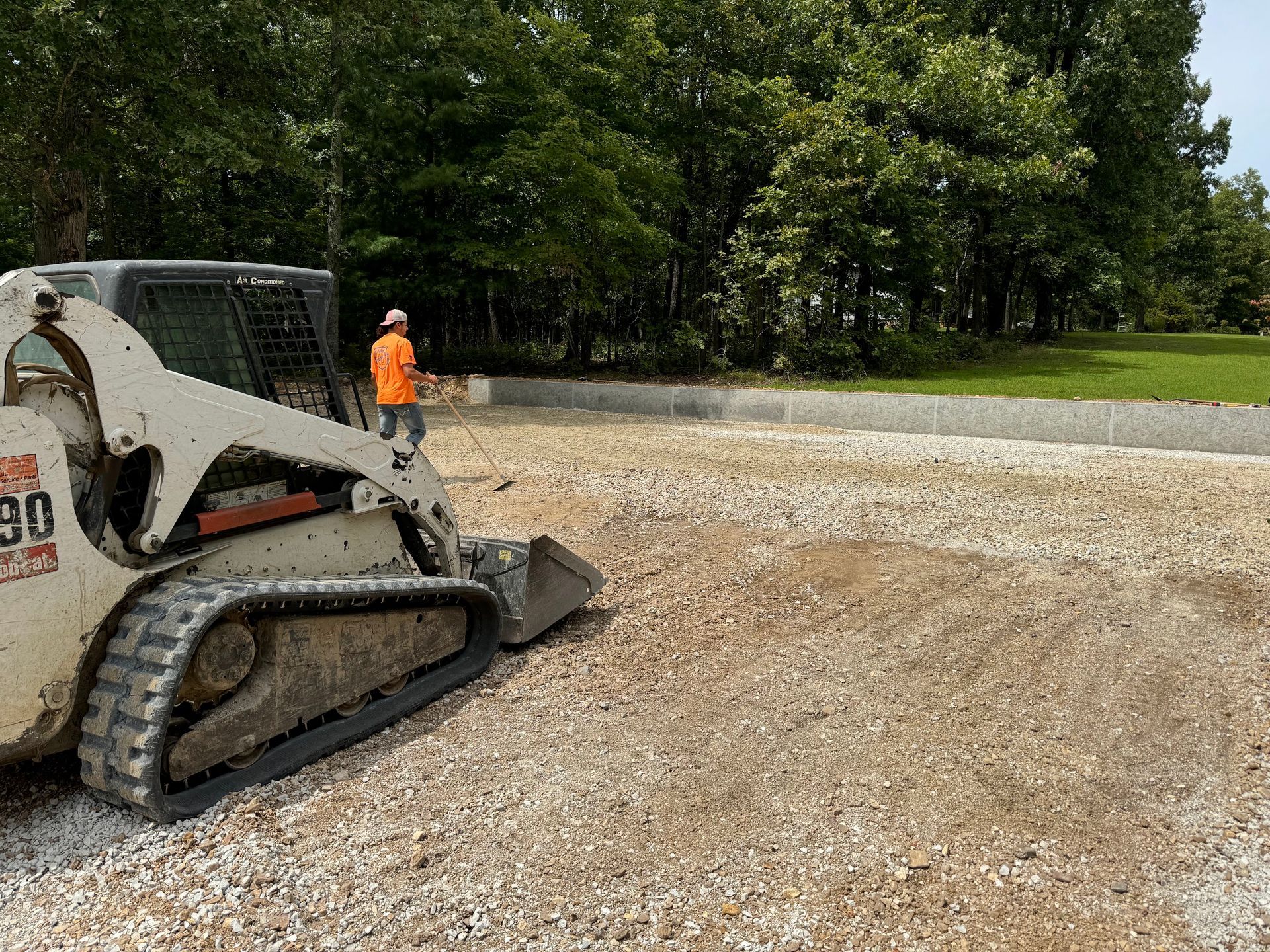 A Bobcat skid steer spreading gravel at a construction site; worker in orange vest, trees in background.