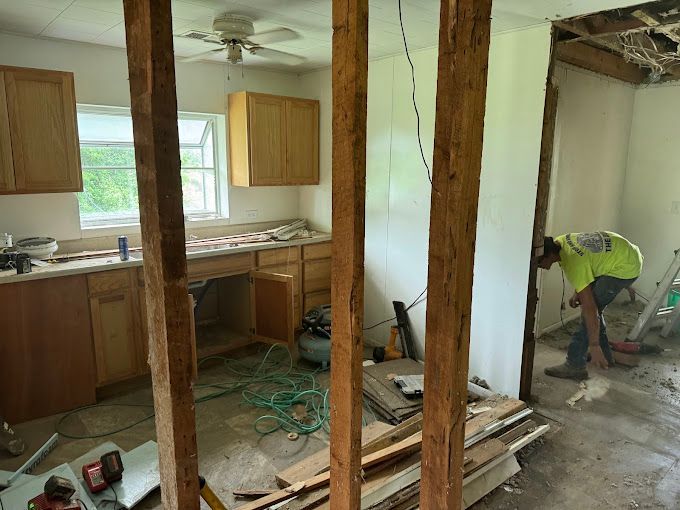 Kitchen under renovation. Worker in neon shirt. Wooden beams, cabinets, window, debris.