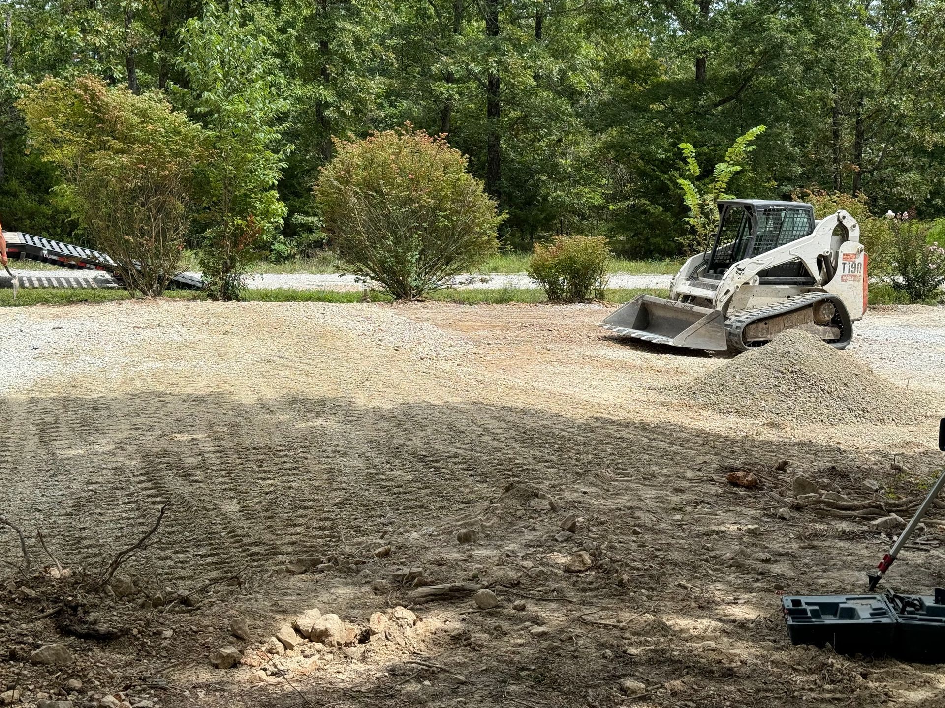 A small white bobcat is spreading gravel in a yard, with trees in the background.