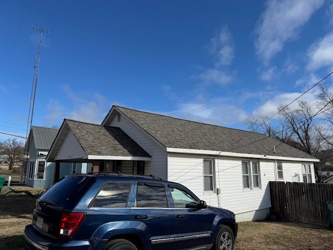 White house with dark gray roof, blue SUV parked in front, blue sky with clouds.