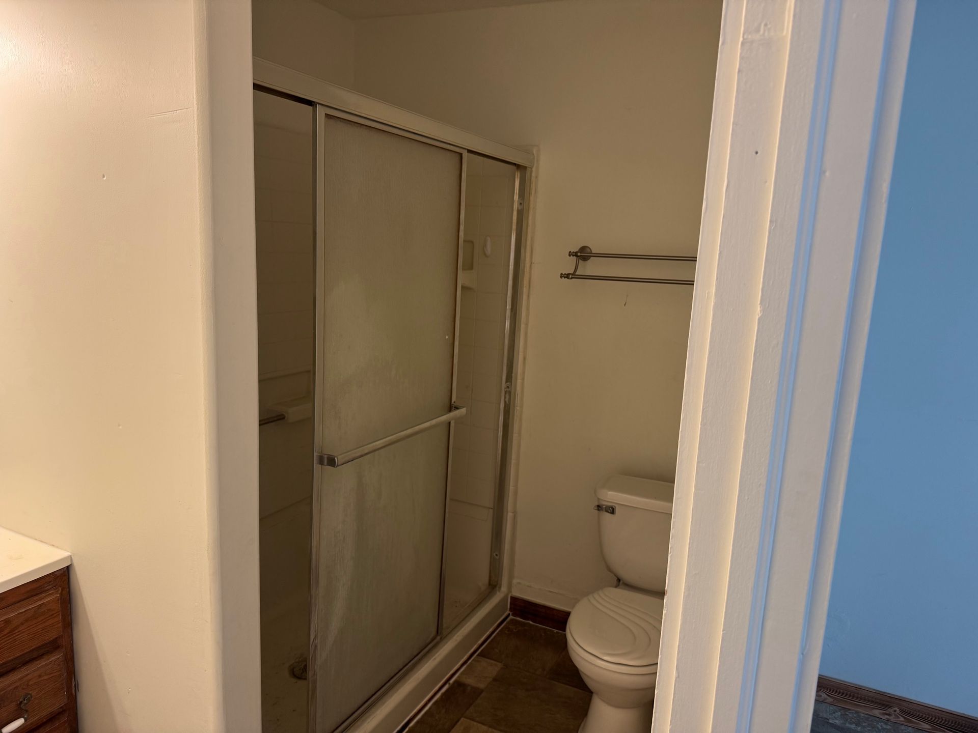 A bathroom with a shower, toilet, and towel rack. White walls, brown flooring.