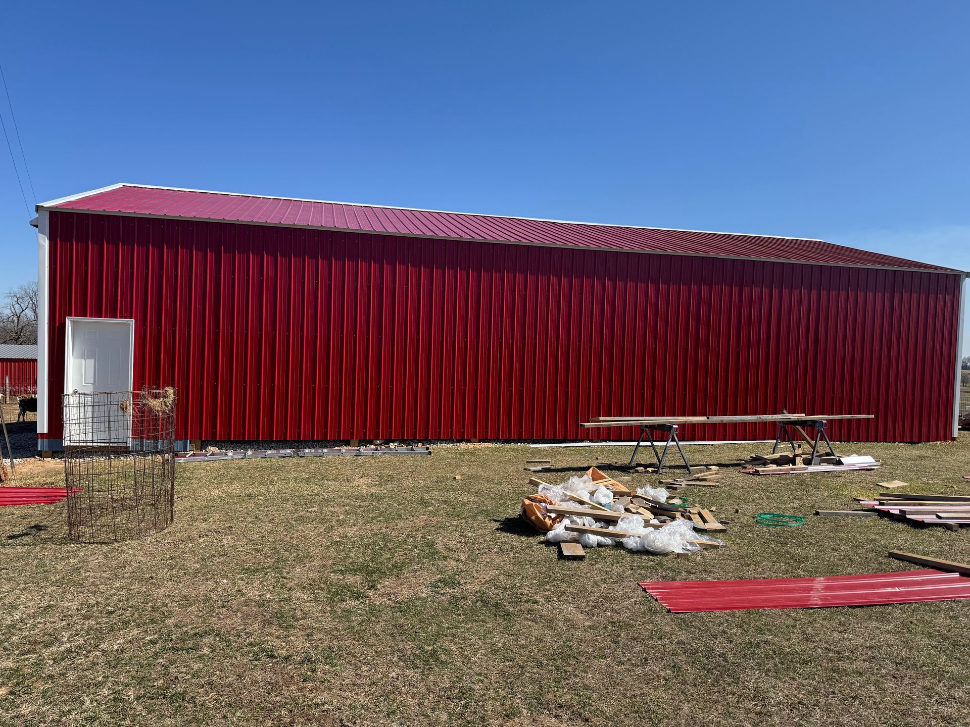 Red metal barn under a blue sky, with a white door, surrounded by construction debris.