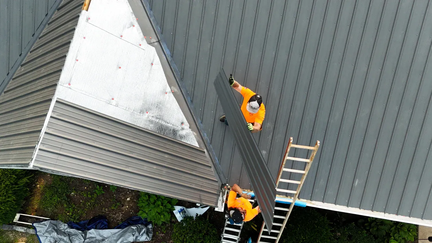 Workers in orange vests installing dark gray metal roof panels on a house.