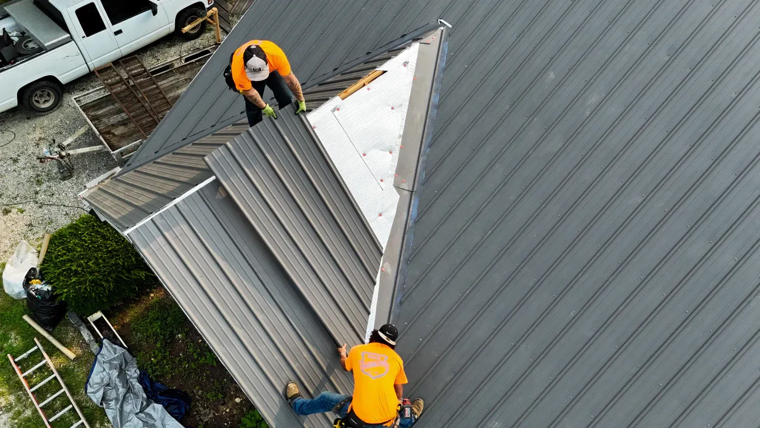 Two roofers in orange shirts install metal roofing on a gray roof.