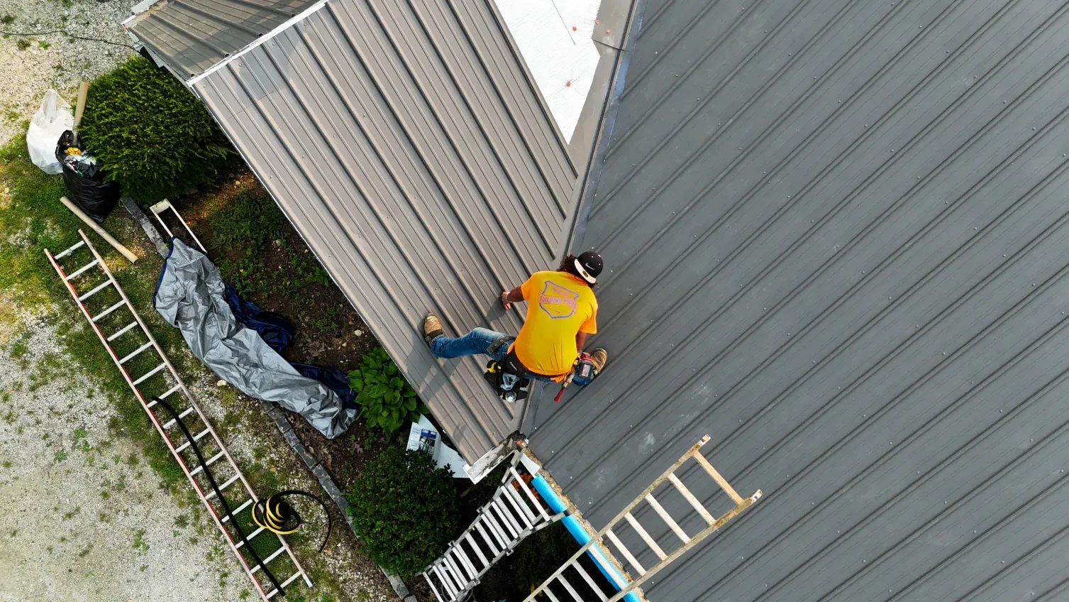 Roofer in an orange vest working on a gray metal roof, ladders nearby.