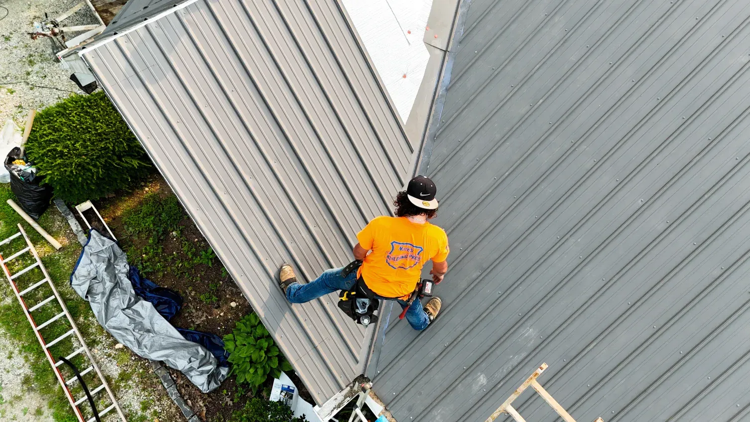 Roofer in orange shirt works on gray metal roof, top-down view.