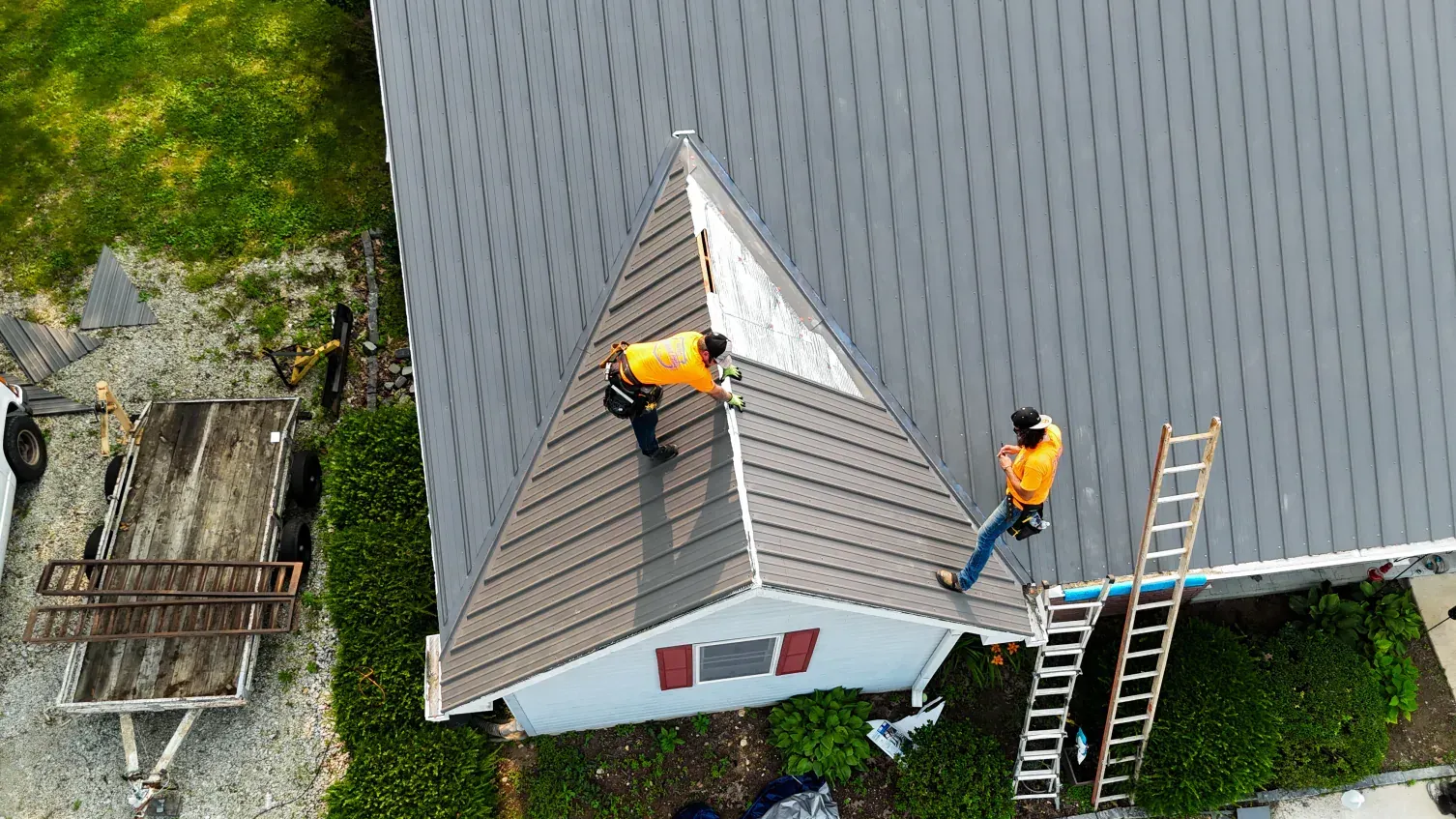 Two roofers in orange vests work on a gray shingle roof, next to a ladder and small white building.