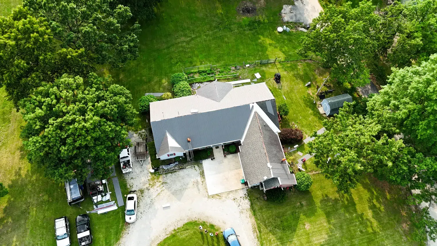Aerial view of a house with gray roof, surrounded by green trees and grass, with vehicles parked nearby.