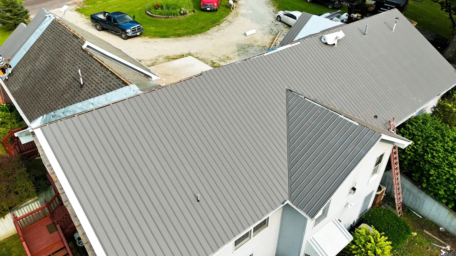 Gray roof on a two-story house with a ladder and a driveway with cars.