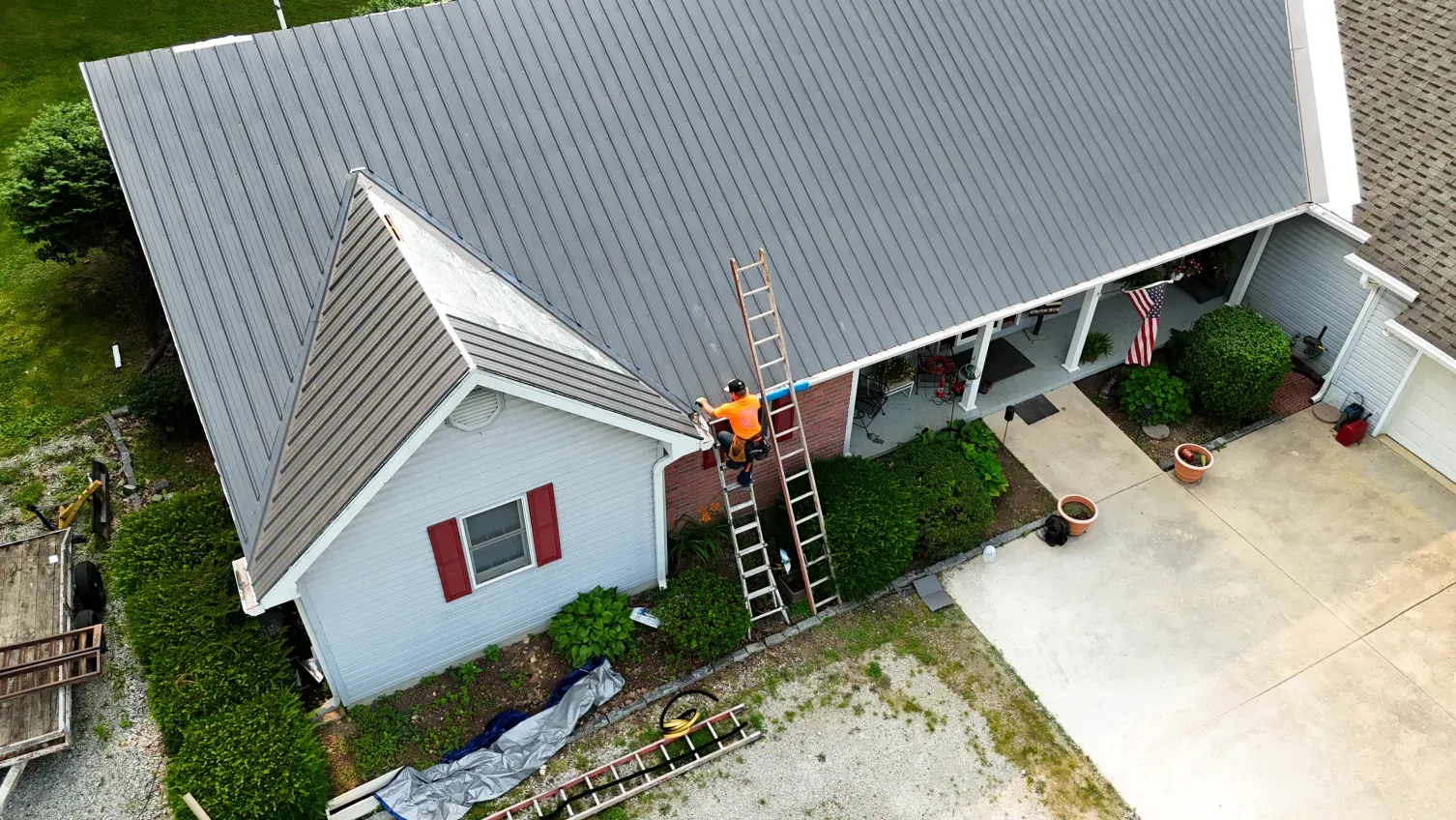 Person on a ladder on gray roof, working on a house with red shutters and a porch.