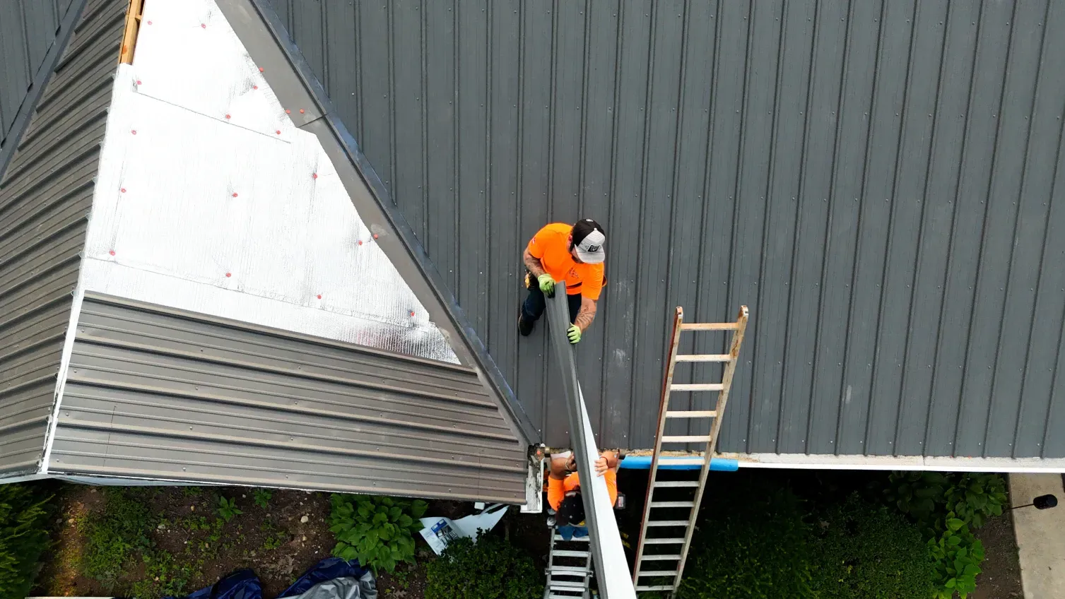 Two roofers on a dark gray roof, one with an orange shirt. Wooden ladder. Green foliage.