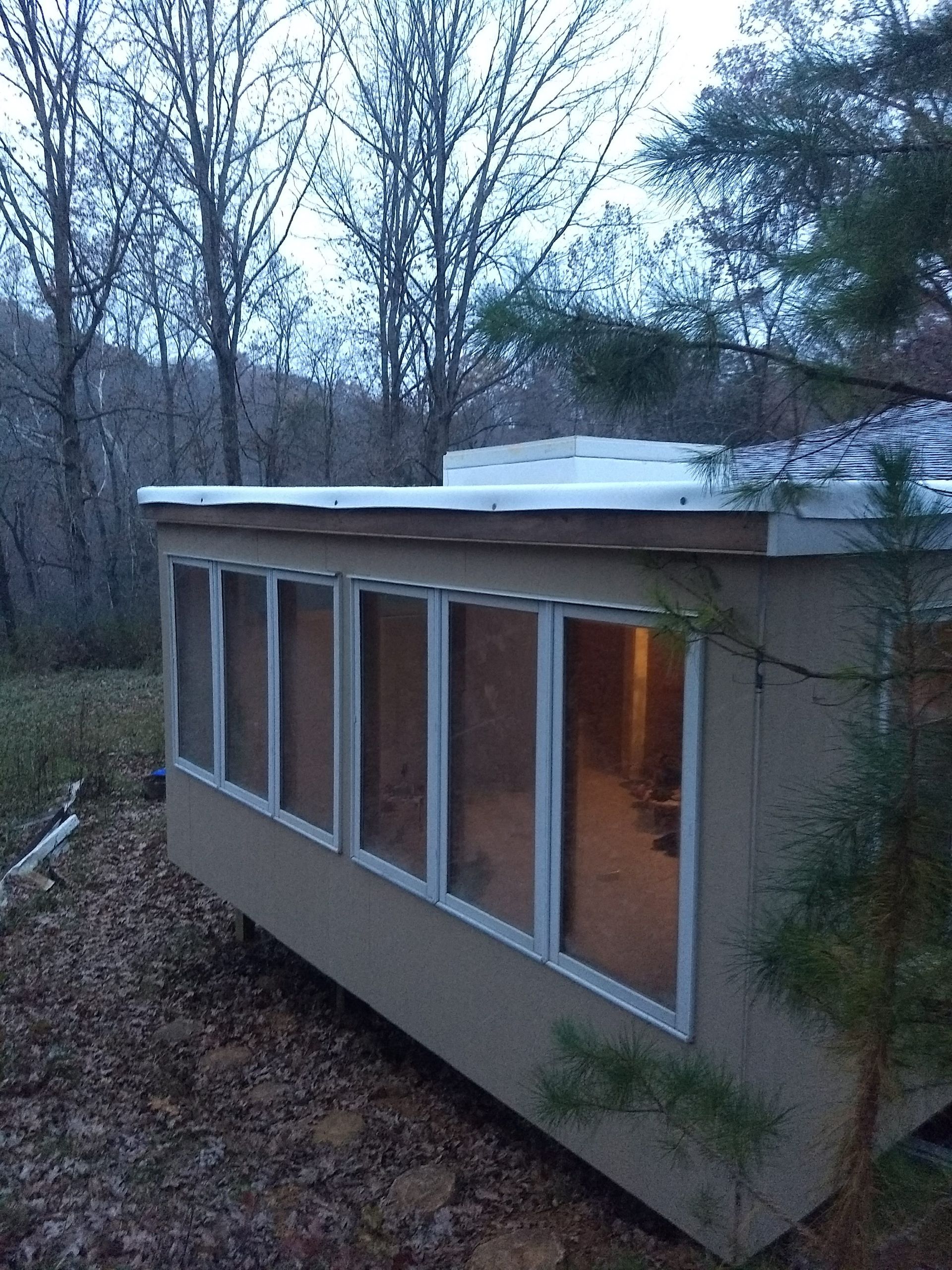 Beige cabin with six tall windows, set in a wooded area, illuminated from within, overcast sky.
