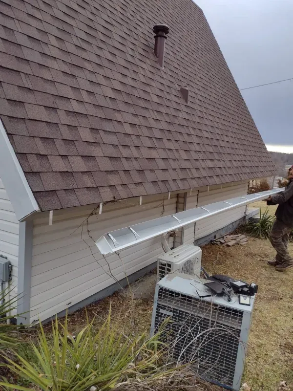 Man installs a long, silver gutter on a white house with brown shingles, with a large air conditioner below.