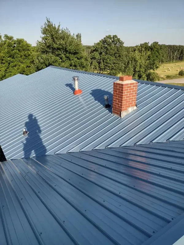 Blue metal roof with chimney and vent; a person's shadow is visible. Trees and open land are in the background.