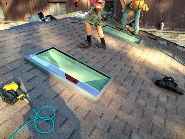 Two skylights being installed on a brown shingled roof by two workers in shorts and work boots.