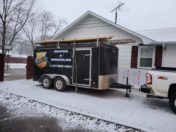 Black roofing trailer parked in the snow, next to a house and truck.  The trailer has a logo and phone number on it.