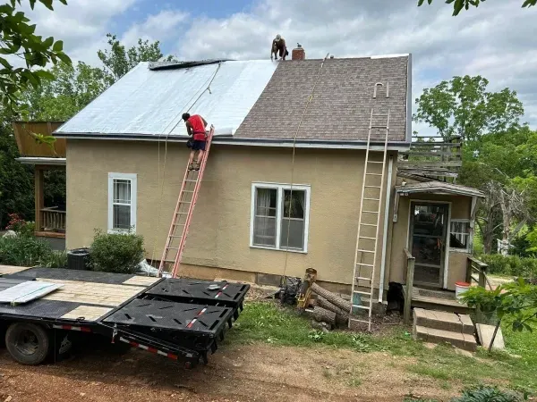 Roofers replacing a roof on a tan house. One person on a ladder, another on the roof, working with silver metal sheets.