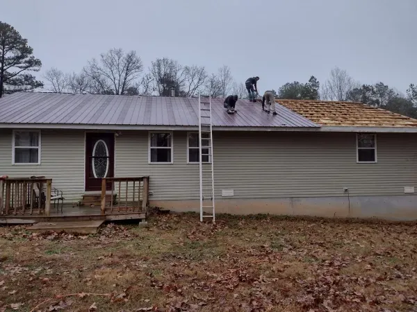 Three workers installing a metal roof on a house. One side complete, one side in progress.