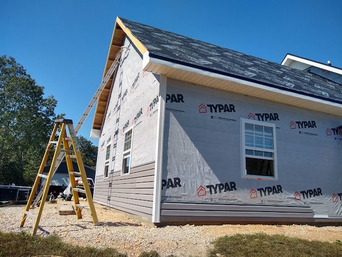 House under construction with siding being installed. Ladder leaning against the side, clear blue sky.