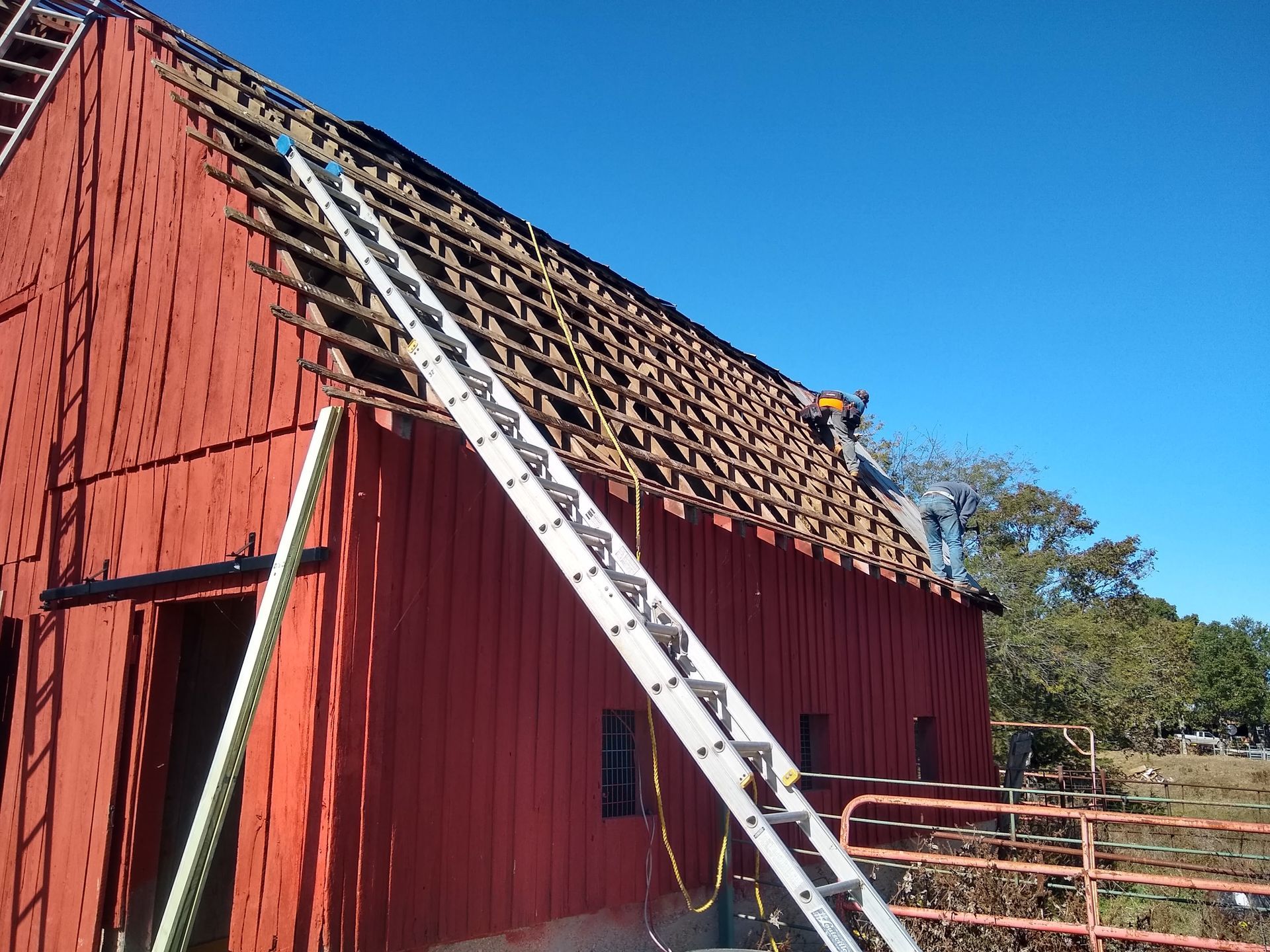 Red barn with roof partially removed; person on ladder working on roof under blue sky.