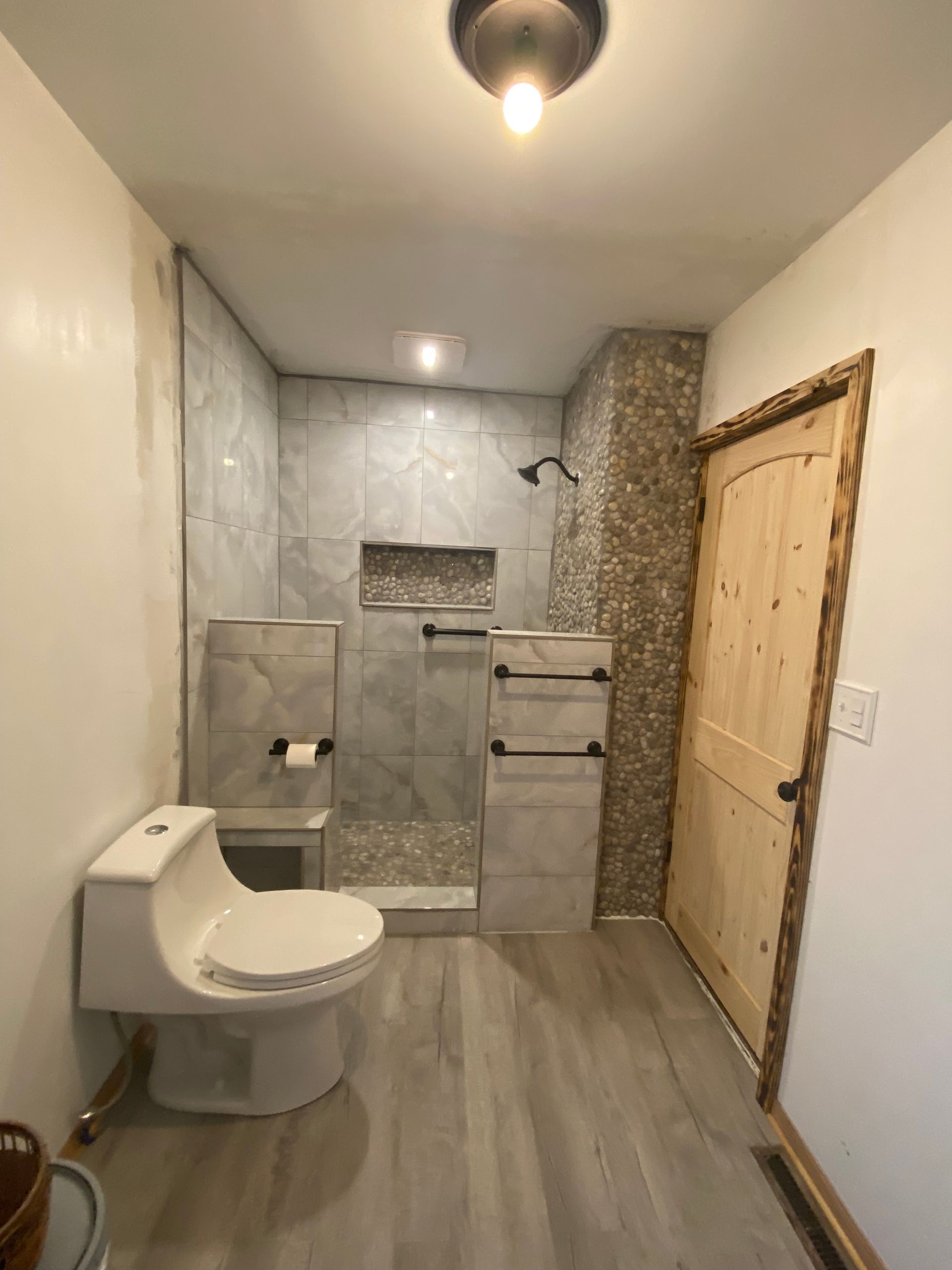 Bathroom with toilet, shower, and wooden door. Gray tile and wood-look flooring.
