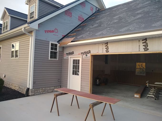 Garage and house under construction; gray siding, pink Typar paper, open garage, white door, concrete.