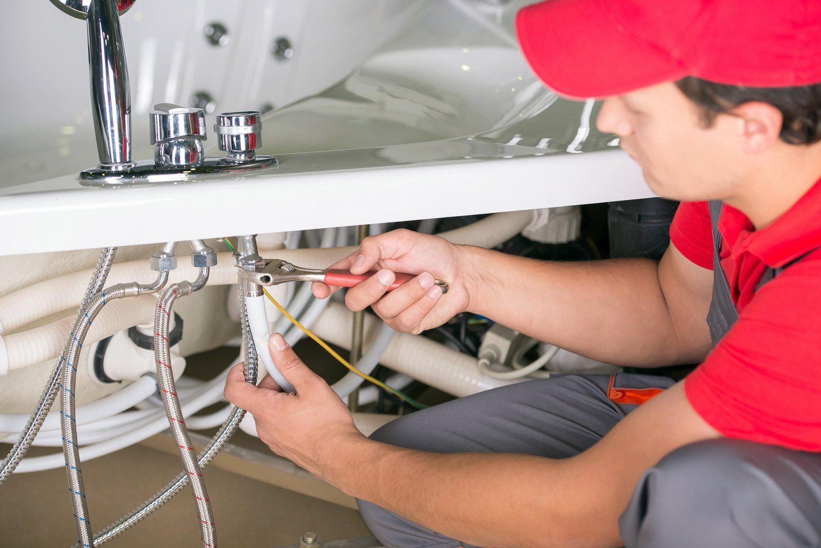 A plumber in a red cap and shirt working under a white bathtub, using a wrench on the pipes.