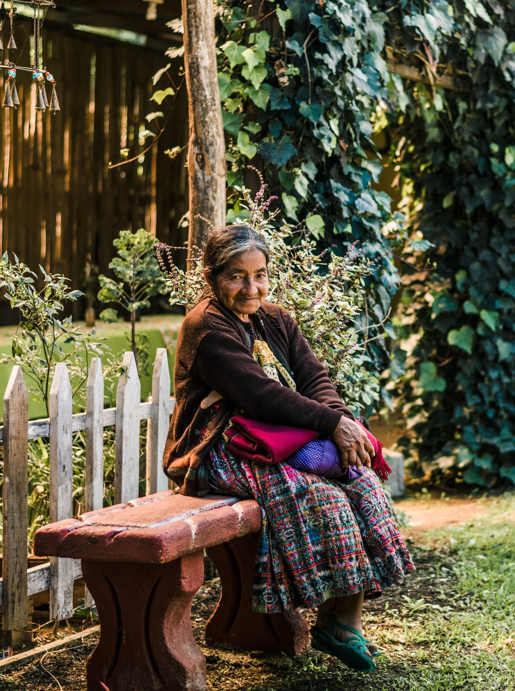 Een vrouw zit glimlachend op een betonnen bankje in een tuin. Ze draagt ​​een rok met een patroon en een bruine trui. Weelderig groen omringt haar.