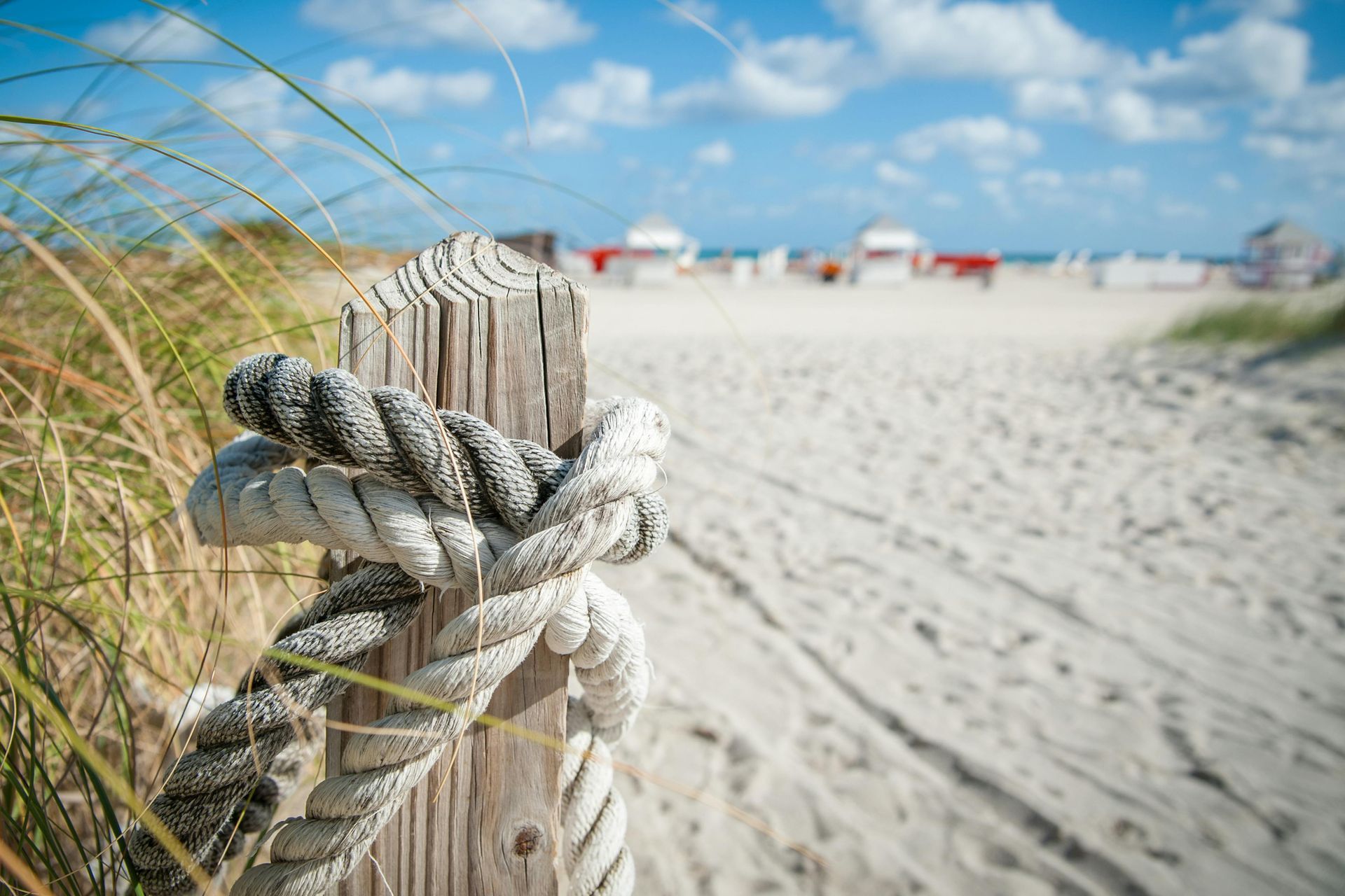 Wooden post with rope, leading to a sandy beach with beach umbrellas under a blue sky.