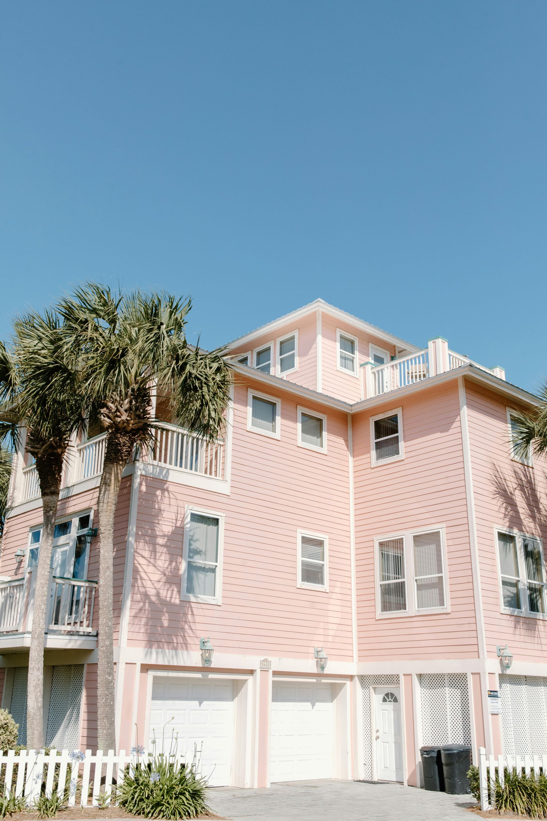 Pink beach house with white trim and garage doors, under a bright blue sky.
