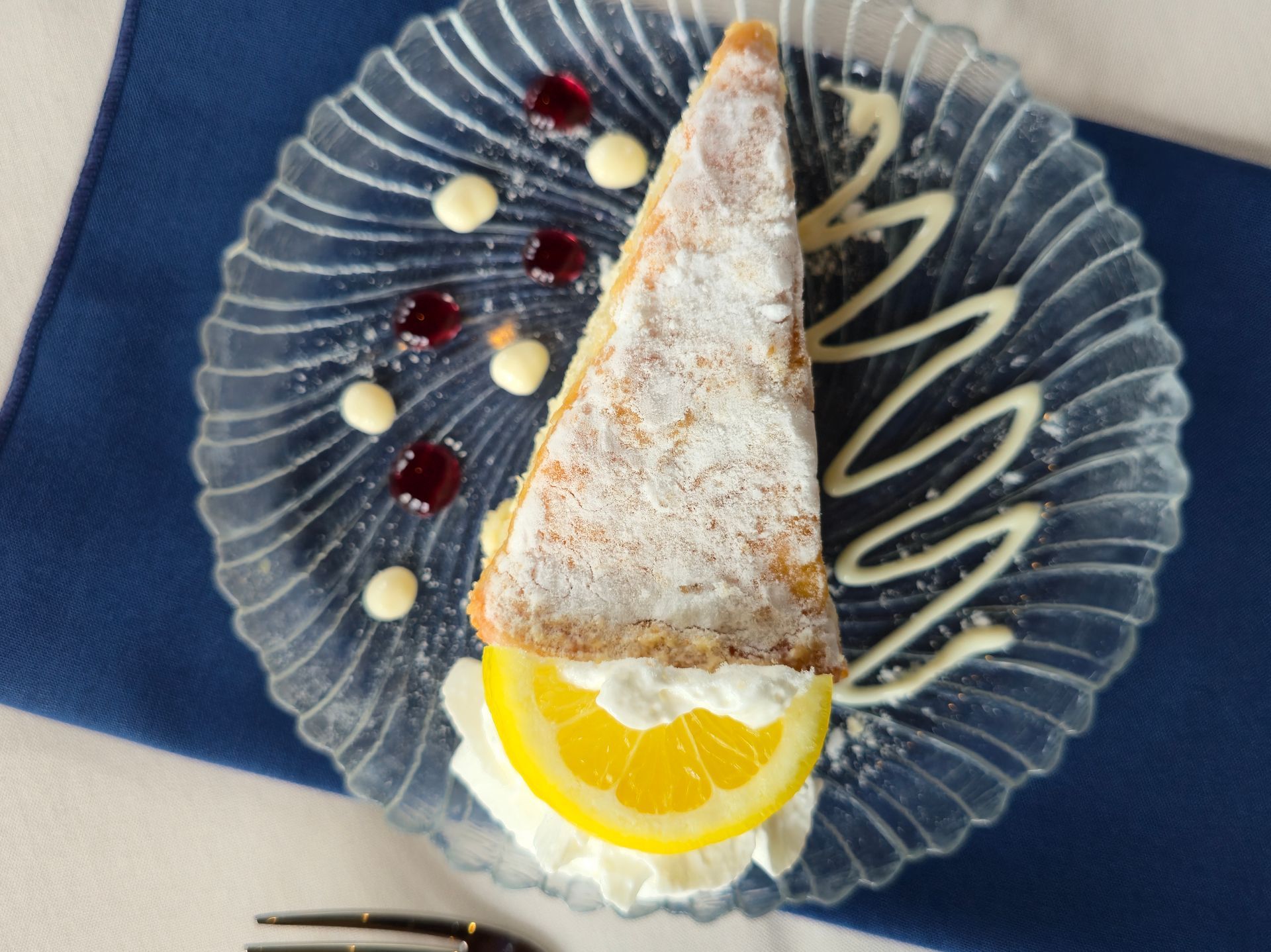 A slice of cake dusted with powdered sugar, garnished with whipped cream and lemon on a glass plate, on a blue napkin.