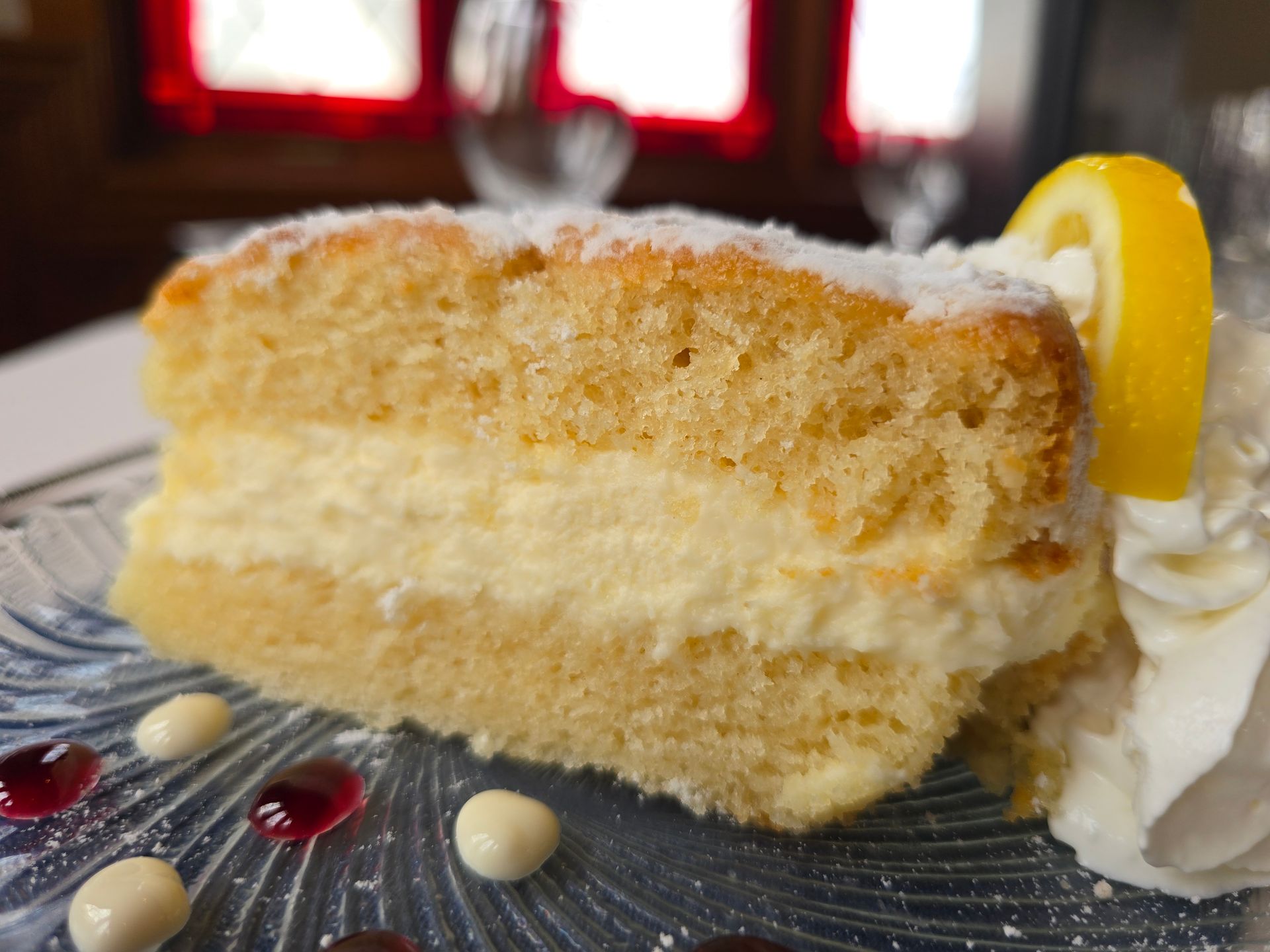 A slice of lemon cream cake on a glass plate, garnished with whipped cream, a lemon slice, and dots of berry sauce.