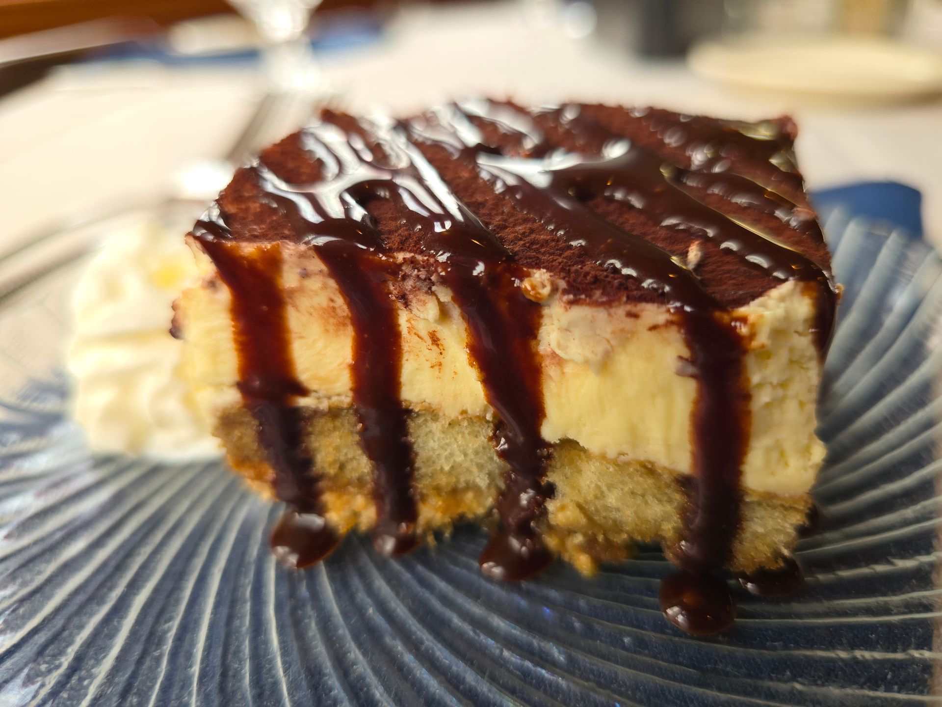 A square slice of tiramisu topped with chocolate drizzle and cocoa powder, served on a blue textured plate with cream.