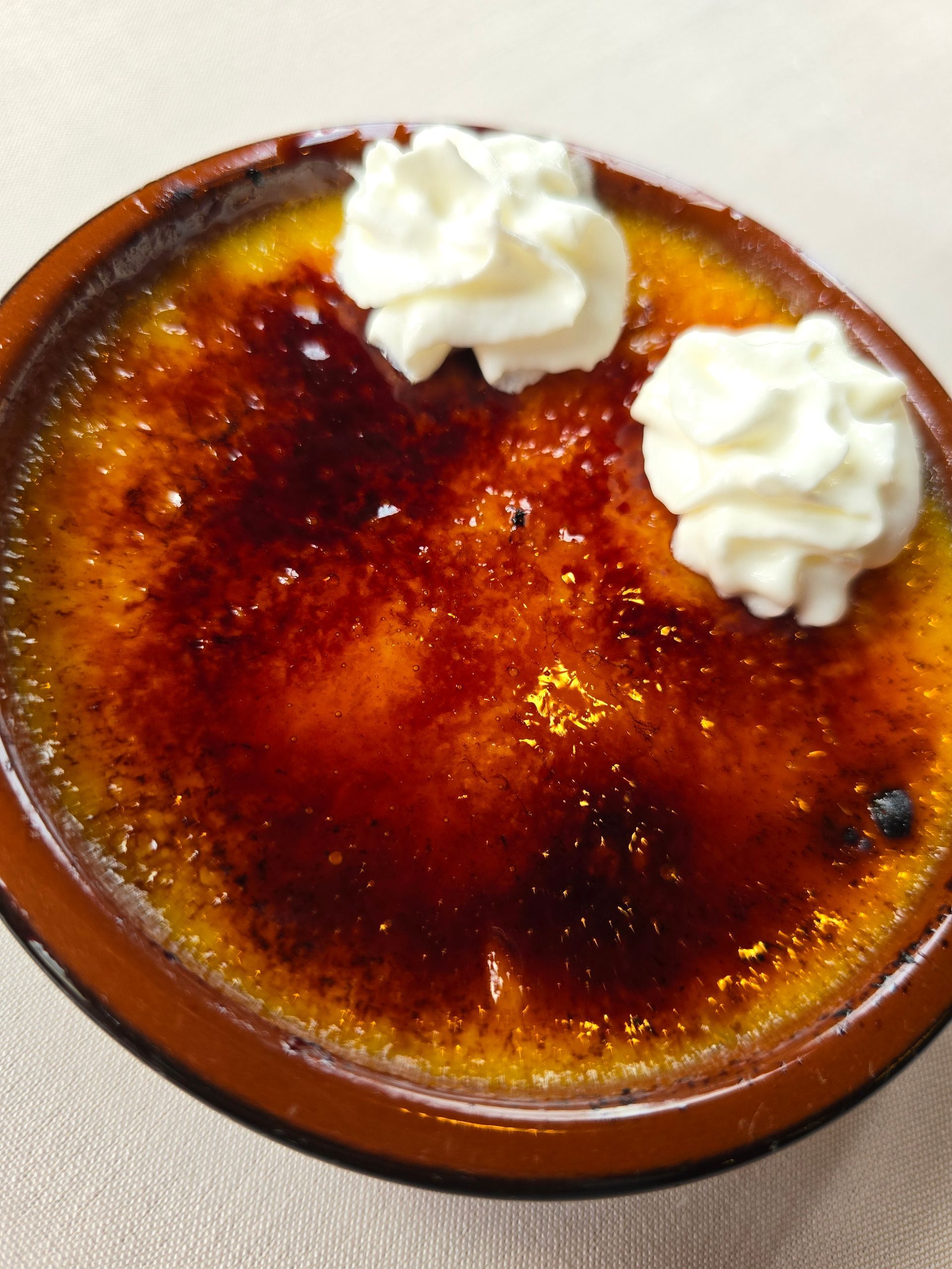 A crème brûlée in a brown ramekin topped with two dollops of whipped cream, set against a neutral background.