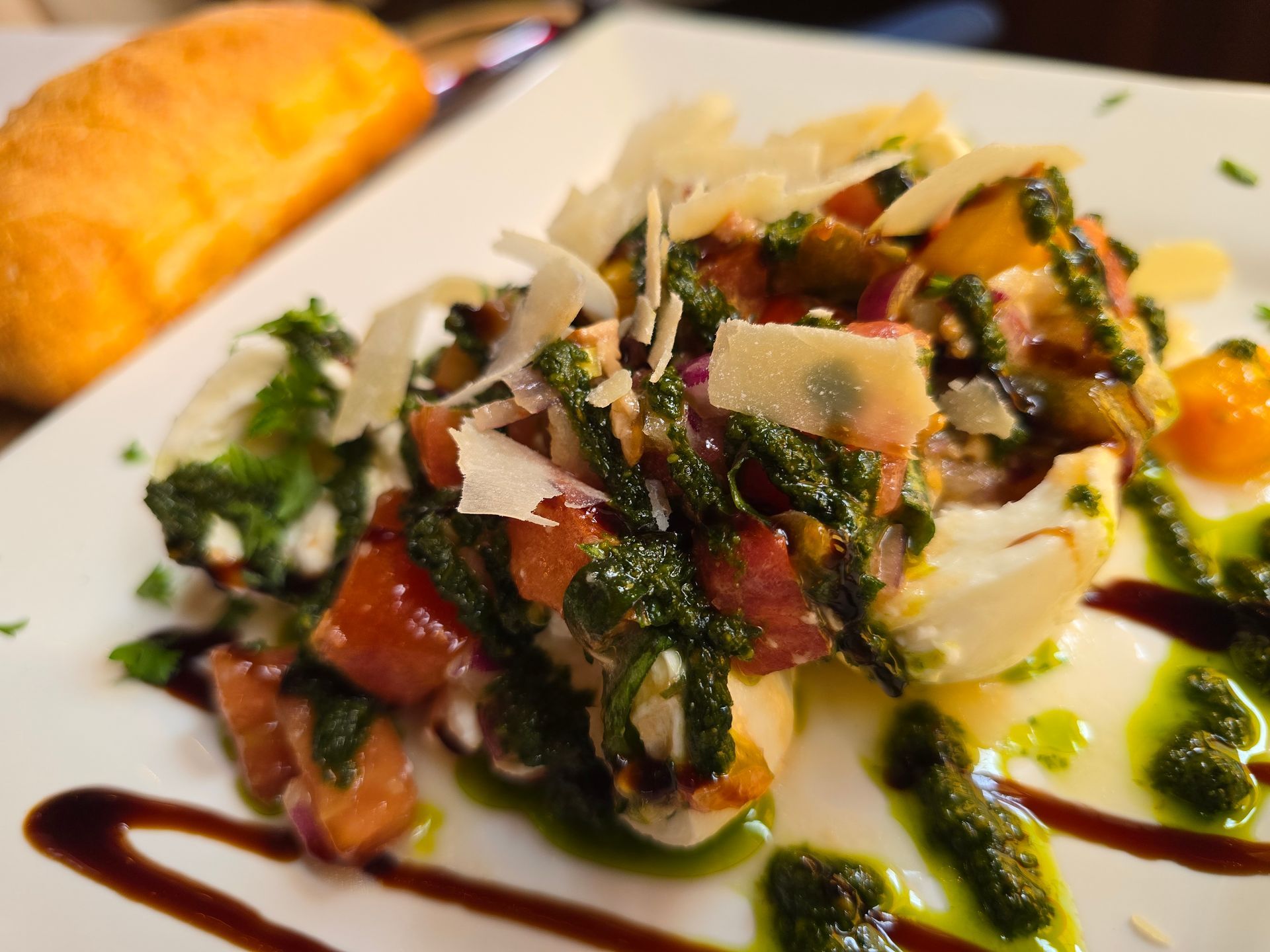 A caprese salad with pesto, balsamic glaze, and parmesan shavings on a white plate next to a piece of bread.