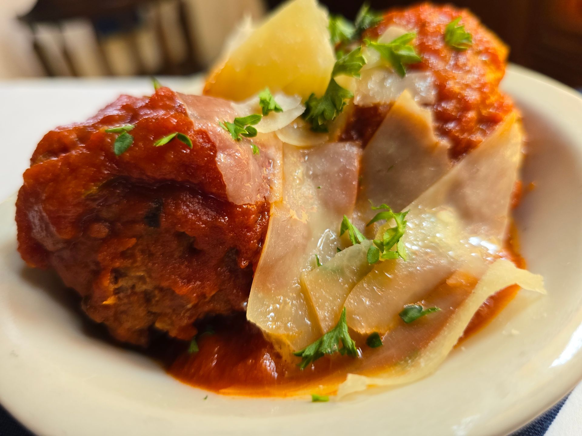 A meatball coated in red tomato sauce, garnished with shaved parmesan cheese and parsley in a white bowl.