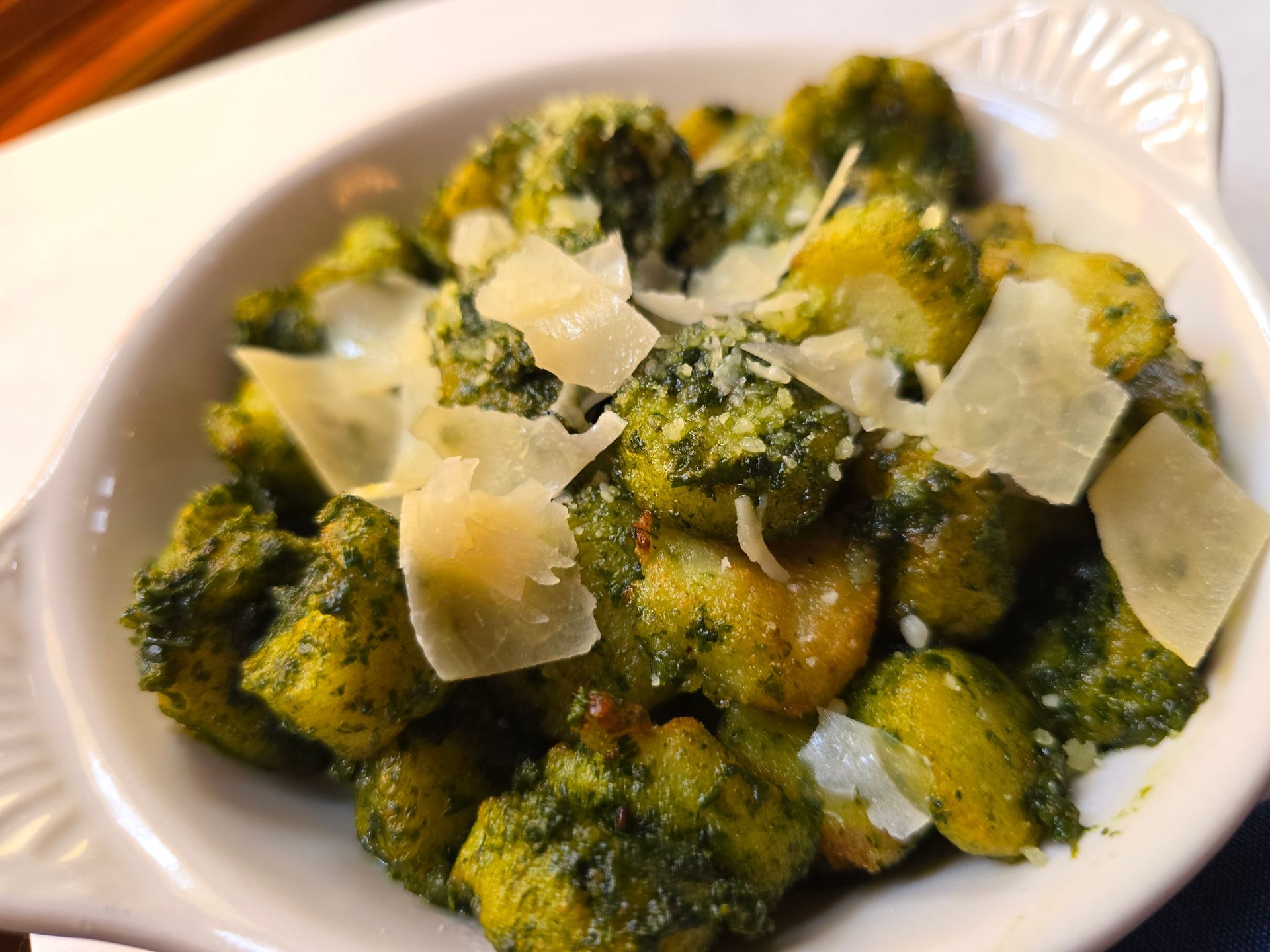 A white serving dish filled with gnocchi tossed in green pesto sauce and topped with shaved parmesan cheese.