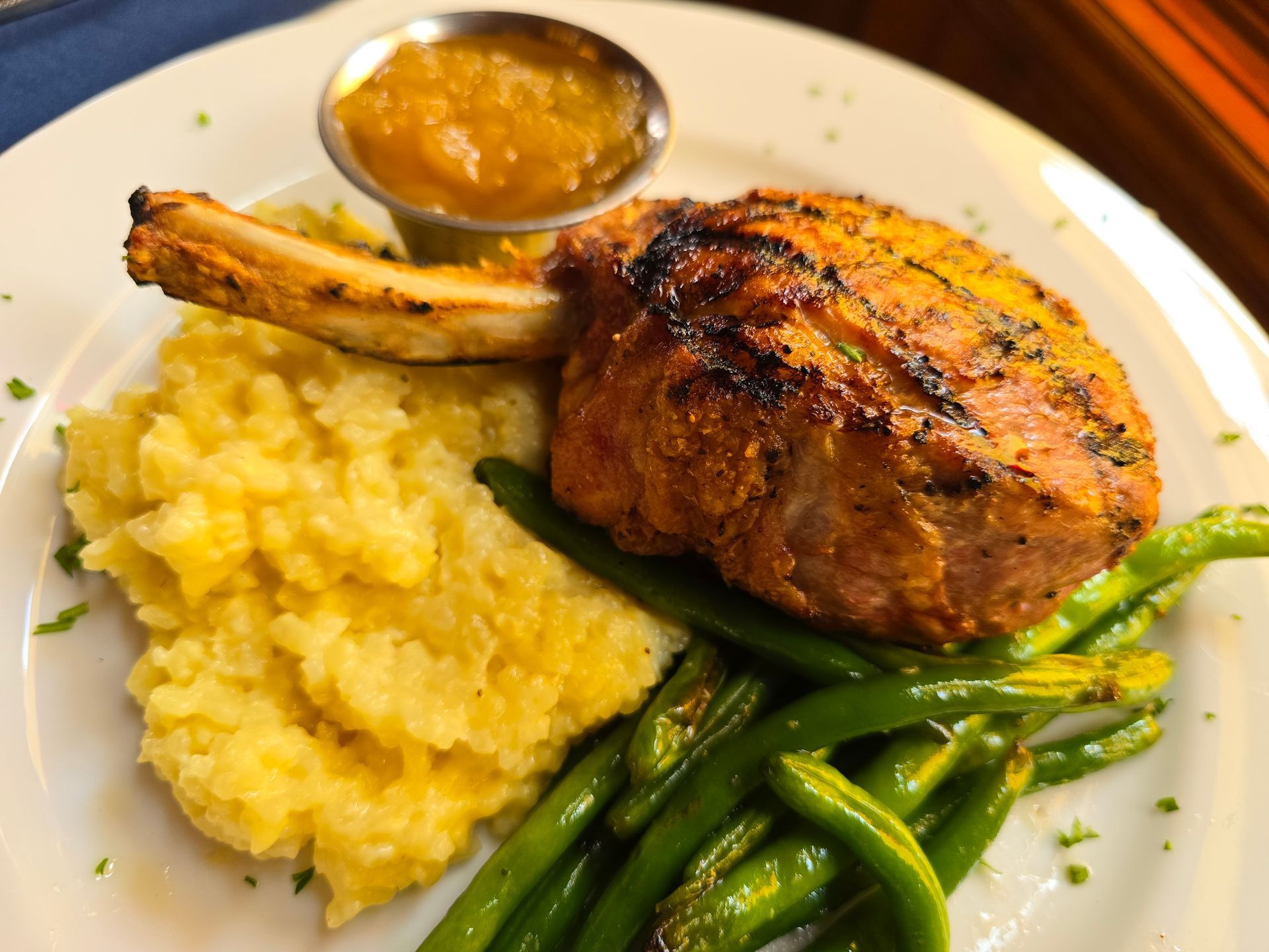A bone-in pork chop served with mashed potatoes, green beans, and a small cup of sauce on a white plate.