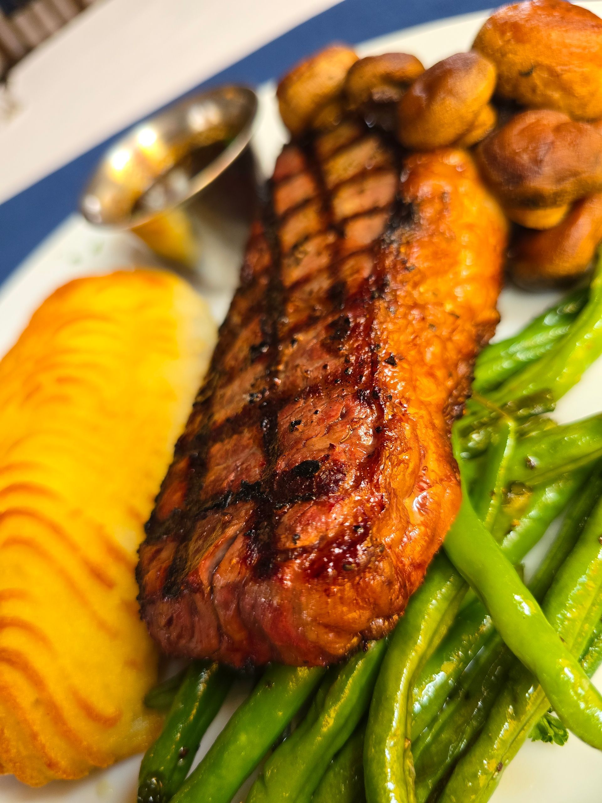 A grilled steak served with sautéed mushrooms, green beans, a side of sauce, and a golden potato cake on a white plate.
