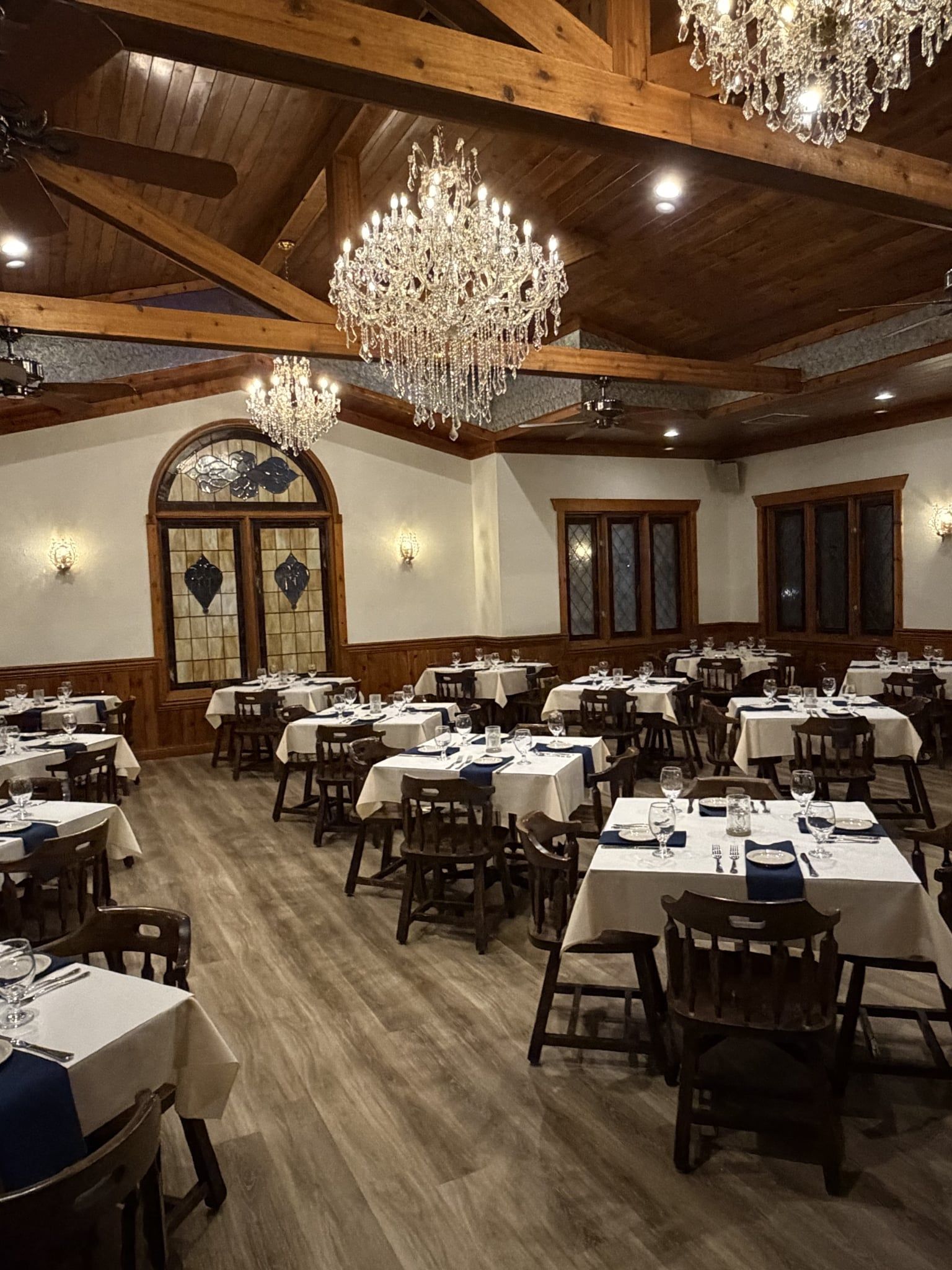 Elegant dining room with chandeliers, wooden beams, tables set for dinner.