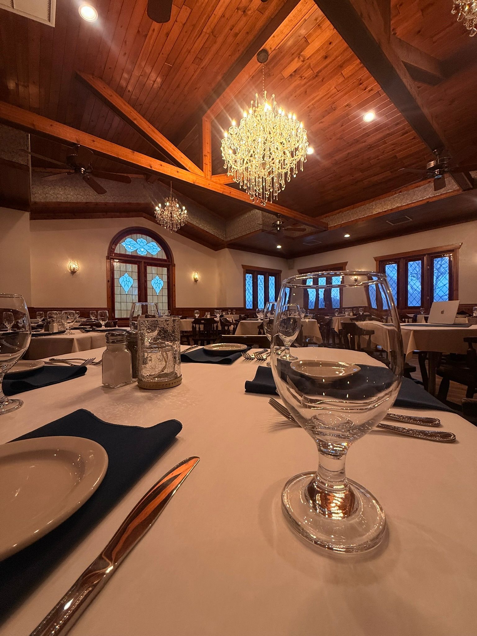 Elegant dining room, tables set with white linens, crystal chandelier hanging from a wooden ceiling.