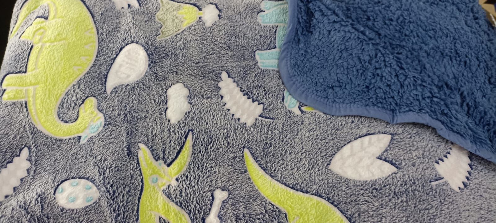Toalla azul sobre una manta gris con estampado de dinosaurio, con detalles en verde y blanco.