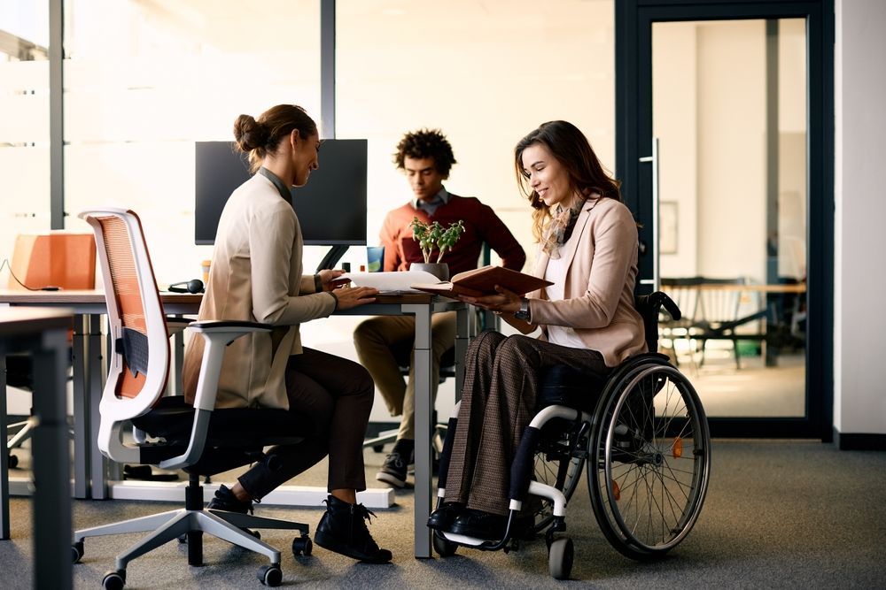 A Woman in a Wheelchair is Sitting at a Table With Two Other People — Better Access Support Services In Ipswich, QLD