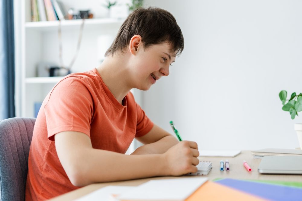 A Young Man is Sitting at a Desk Writing on a Piece of Paper — Better Access Support Services In Logan, QLD