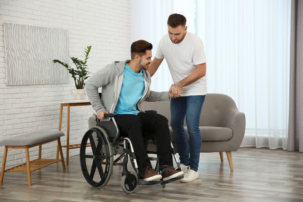 Man helping another man out of a wheelchair in a bright living room.