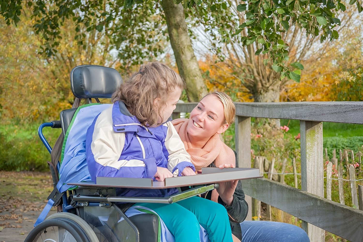 Woman Smiles At Child With Disability In A Wheelchair Outdoors