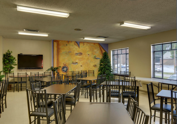 Empty dining hall with tables and chairs, wall art, and large windows letting in daylight Empty dining hall with tables and chairs, wall art, and large windows letting in daylight