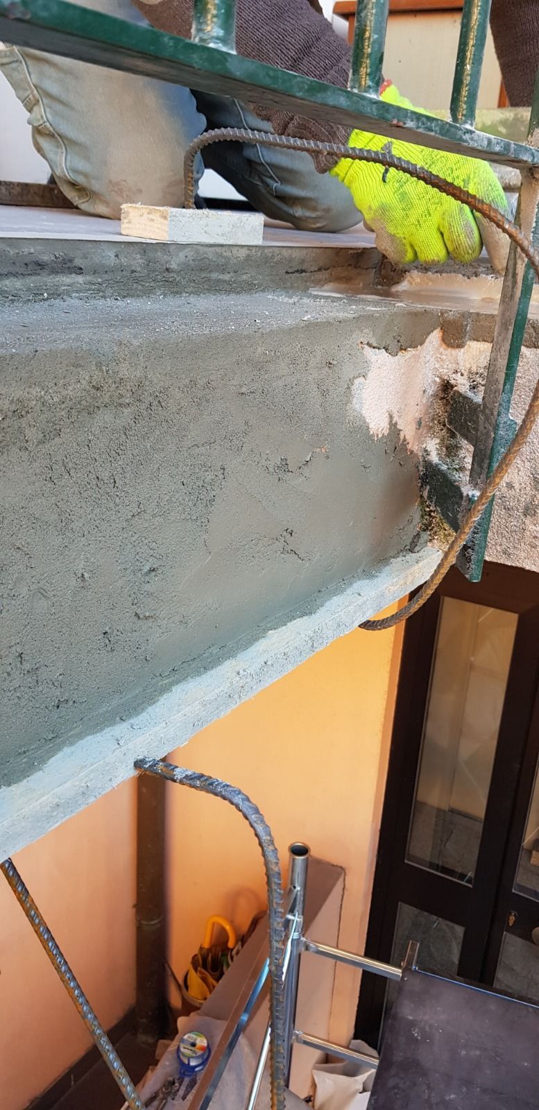 Un balcone color salmone con ringhiera in vetro, tenda da sole e una finestra parzialmente visibile nella parte inferiore.