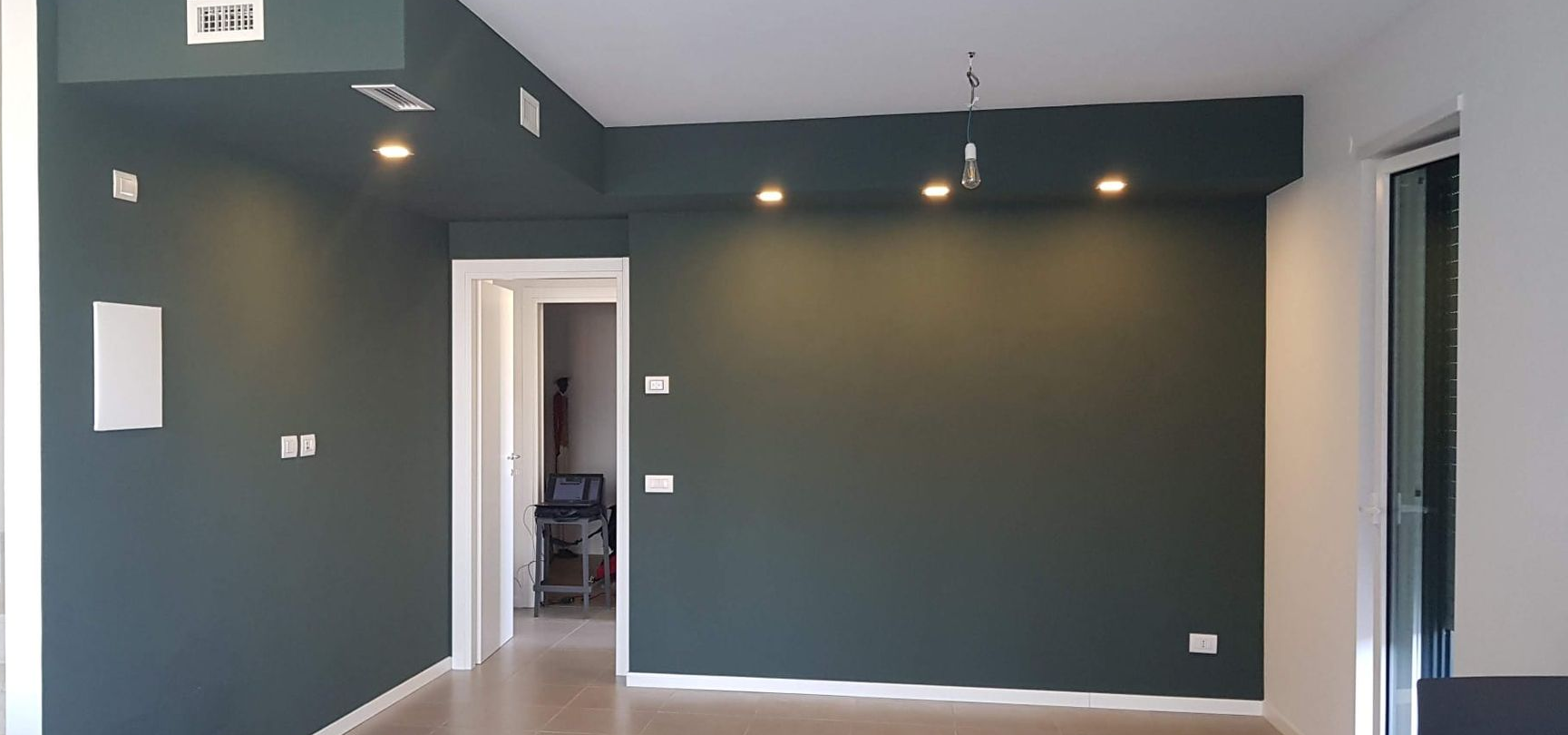 Una stanza vuota con pareti verde scuro e soffitto bianco. Sono visibili una porta e delle luci.